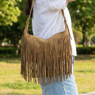 A woman wearing a white shirt and light blue jeans carries a brown suede fringe shoulder bag in an outdoor park setting.