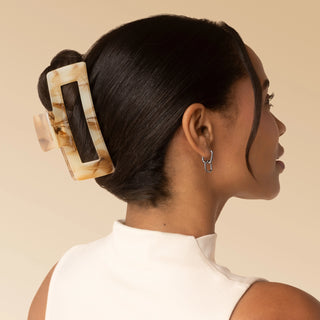 A woman shown in side profile with her hair styled in a low bun, wearing a large rectangular tortoiseshell hair claw clip. The background is a solid, warm beige studio backdrop.