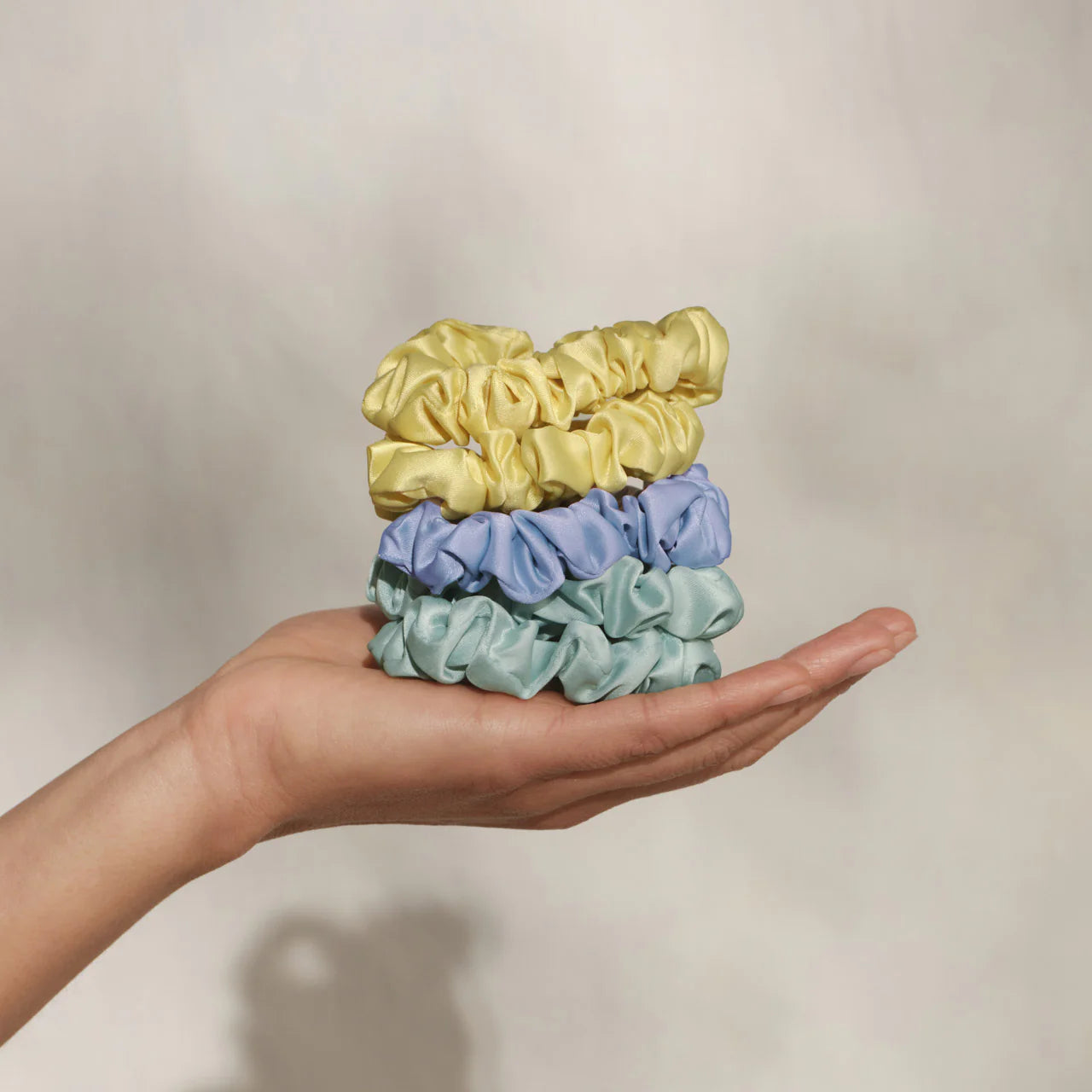 Hand holding four scrunchies in yellow, blue, and green on a neutral background
