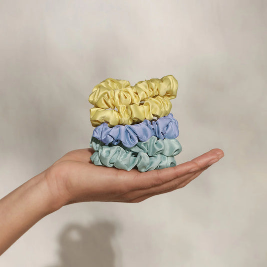 Hand holding four scrunchies in yellow, blue, and green on a neutral background
