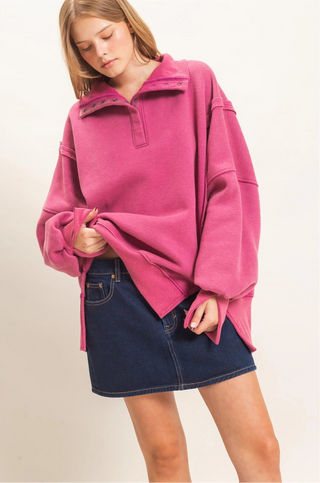 A woman wearing an oversized bright pink mock-neck sweatshirt and a dark blue denim mini skirt, standing against a plain off-white background.