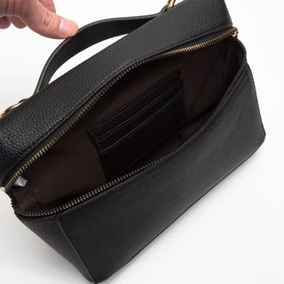A black pebbled leather cosmetic bag shown open, with a hand holding the handle. The bag features gold-tone zippers, a black interior card slot, and a dark brown lining.