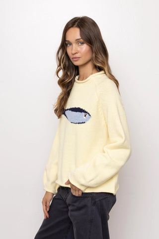 A woman modeling a pale yellow fuzzy crewneck sweatshirt with a sequined fish graphic on the chest, paired with dark gray pants, against a plain white studio background.