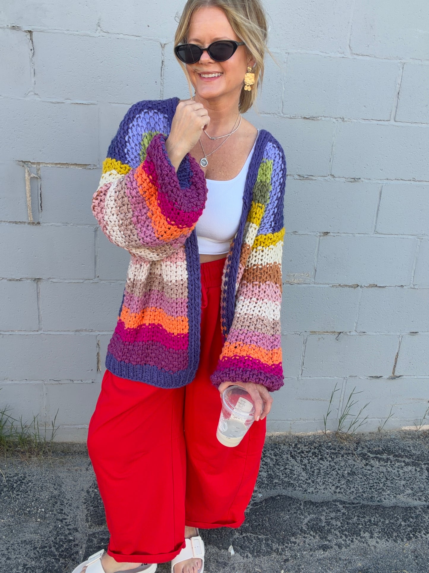 Woman wearing a colorful crocheted cardigan and red pants against a gray brick wall.