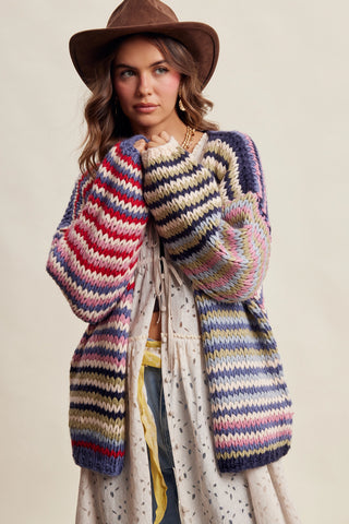 A woman wearing a chunky knit cardigan with horizontal multicolor stripes in navy, pink, red, yellow, and cream, styled with a brown wide-brim hat and a cream dress against a warm beige background.