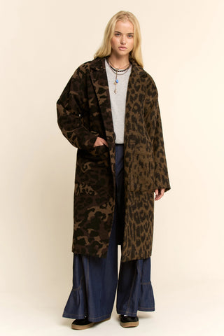 A blonde model stands against a plain beige studio background wearing a long brown and black leopard-print coat over a light gray top and dark blue wide-leg pants.