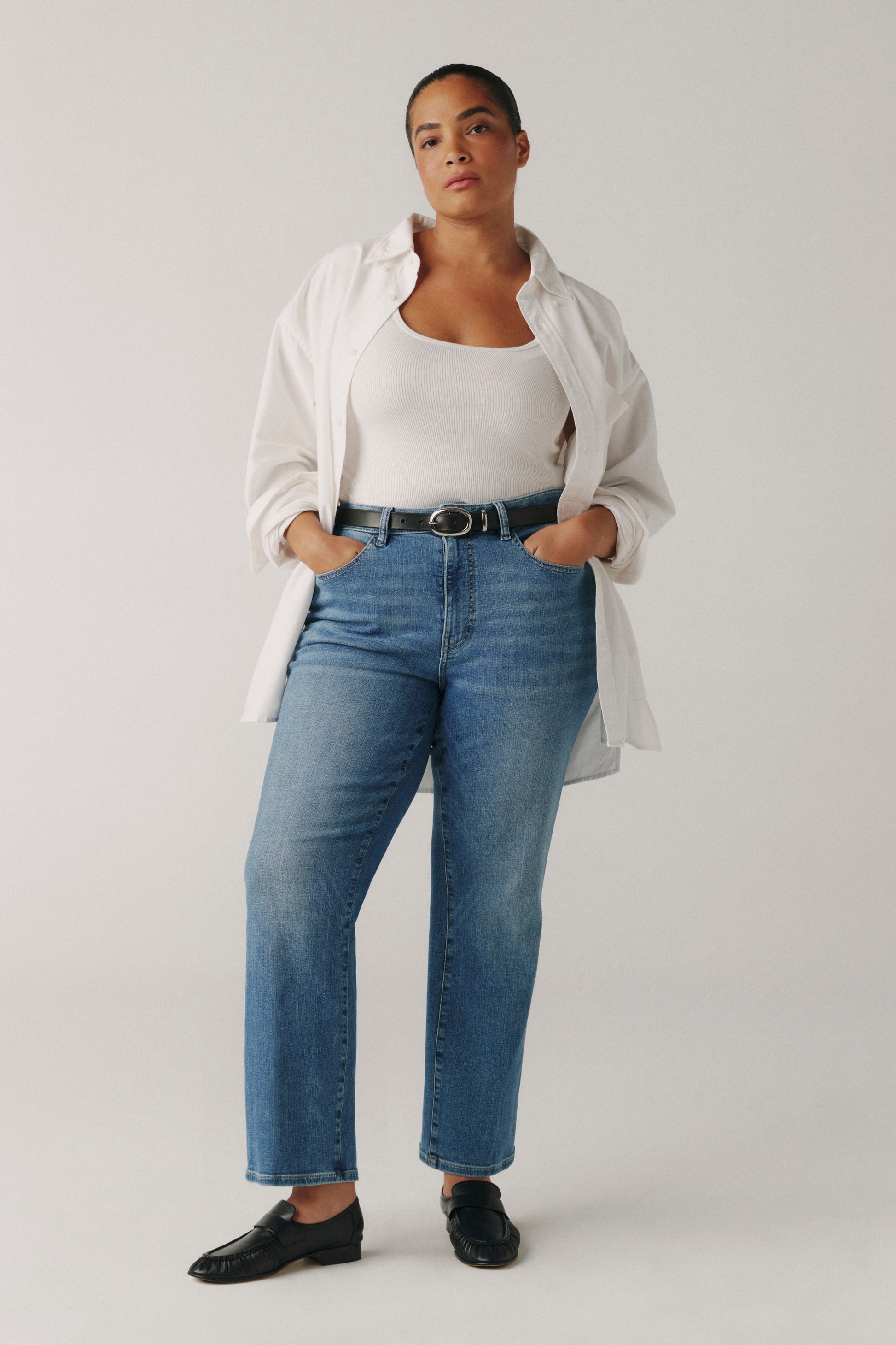 Person wearing a white shirt, white top, blue jeans, and black shoes on a plain background