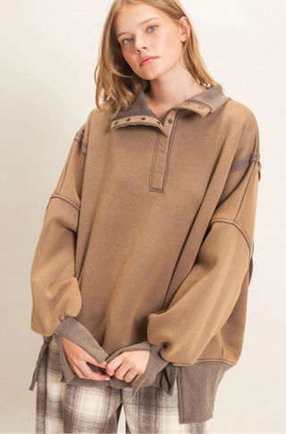 A woman wearing an oversized taupe-brown pullover sweatshirt with contrasting darker brown trim and a half-placket collar, paired with plaid pants, against a plain off-white background.