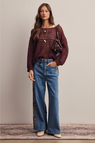 A woman stands in a studio wearing a burgundy ribbed knit long-sleeve top, blue wide-leg jeans with floral embroidery on the lower left leg, and beige pointed shoes. She is carrying a dark brown shoulder bag against a plain beige background.