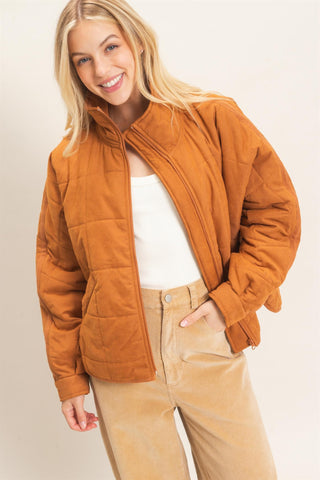 A smiling blonde woman wearing a cropped, quilted zip-up jacket in a warm camel-brown color, styled with a white top and tan pants against a plain light background.