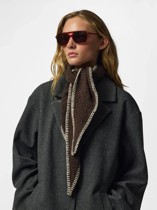 A woman wearing a dark gray wool coat, a brown knit scarf with cream-colored chain trim, and dark red sunglasses, photographed against a plain light gray background.