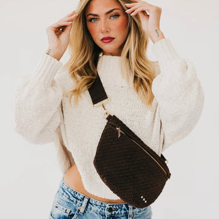 A woman wearing a cream knit sweater and blue jeans models a dark brown textured crossbody bag with gold hardware against a plain white background.