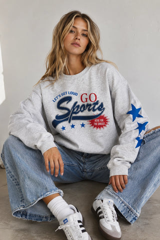 A woman sits cross-legged wearing a light heather gray sweatshirt with a patriotic 'GO Sports' graphic in red, white, and blue, paired with blue jeans and white sneakers.
