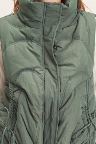 Close-up of a sage green puffer vest with a high stand-up collar, front zipper, and side drawstring details.
