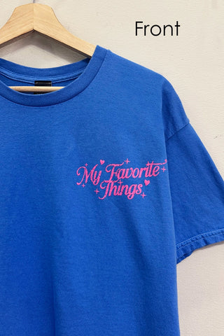 A blue short-sleeve crewneck t-shirt hanging on a wooden hanger against a light beige wall. The shirt features the phrase 'My Favorite Things' printed in pink cursive script with small pink hearts and stars on the left chest.
