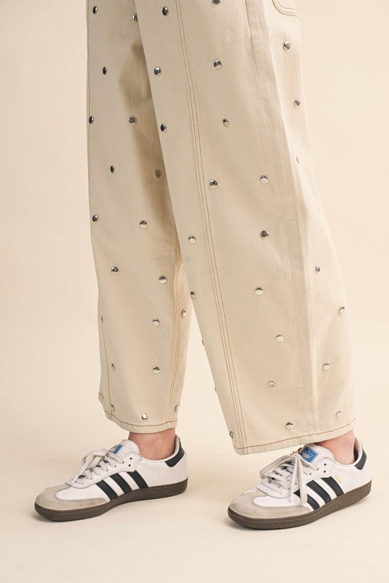 Person wearing beige pants with black embellishments and Adidas sneakers on a beige background