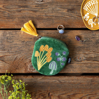 A green velvet coin purse with embroidered mushrooms and a yellow tassel, styled on a rustic wooden background with jewelry and flowers.