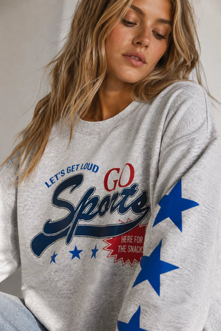 A woman wearing a light heather gray sweatshirt with a bold graphic print that reads 'LET'S GET LOUD GO Sports HERE FOR THE SNACK' in navy and red, with large blue stars printed down the sleeve.