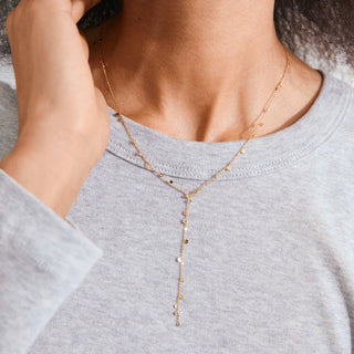 A close-up of a model wearing a delicate gold Y-necklace with small dangling charms, paired with a light gray crewneck sweatshirt.