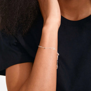 A delicate silver chain bracelet with small charms worn on a person's wrist, shown in a close-up shot against a black t-shirt.