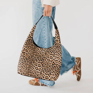 A person wearing light blue jeans and brown lace-up shoes carries a large leopard-print shoulder bag with black trim and a black handle against a clean white background.