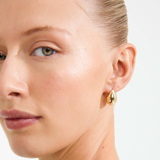 A close-up of a model's ear wearing a small, polished gold teardrop-shaped stud earring against a clean white background.