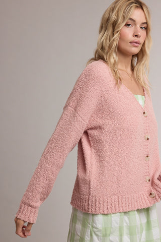 A woman wearing a pink fuzzy button-front cardigan over a light green and white checkered dress, photographed against a plain light gray studio background.
