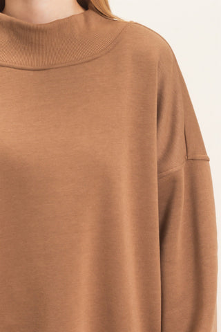 Close-up of a person wearing a soft camel-brown knit top with a ribbed mock neck and long sleeves, against a plain light background.
