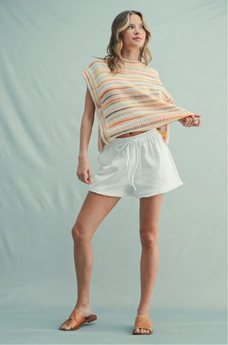 A woman modeling a knit sleeveless top with horizontal stripes in cream, orange, and yellow, paired with white shorts and tan slide sandals, against a light blue-green studio backdrop.