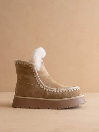 A beige suede ankle boot with white faux-fur trim and a chunky platform sole, photographed against a warm beige studio background.