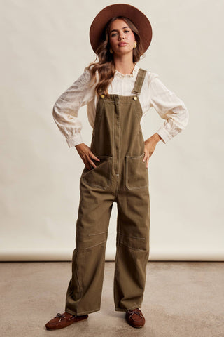 A woman modeling wide-leg olive green canvas overalls over a white ruffled blouse, wearing a brown wide-brim hat and brown shoes, against a neutral beige studio backdrop.