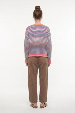 A model viewed from behind wearing a striped knit sweater with horizontal bands of purple, pink, and blue, paired with brown corduroy trousers and tan sandals, against a plain white studio background.