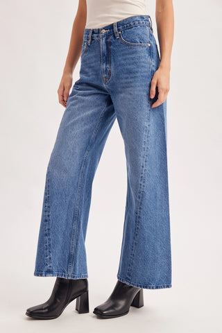 A model wearing a pair of high-waisted, wide-leg blue denim jeans with black leather block-heel boots, shown from the waist down against a plain white studio background.