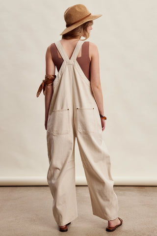 A woman shown from the back wearing a light beige wide-leg overall jumpsuit with brown straps, a brown sleeveless top, a brown hat, and sandals, standing against a neutral cream backdrop.