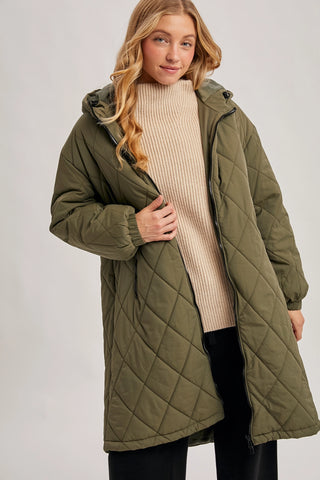 A woman wearing a long olive green quilted puffer coat over a beige ribbed turtleneck sweater and black pants, photographed against a plain light gray studio background.