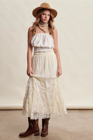 A woman modeling a bohemian-style cream two-piece set consisting of a strapless ruffled crop top and a matching lace maxi skirt, styled with a tan hat, beige choker, and brown cowboy boots against a neutral cream backdrop.