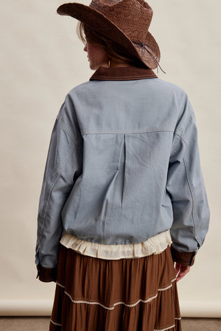 A person shown from the back wearing a light blue cropped jacket with brown trim, a brown hat, and a brown skirt against a plain beige studio background.
