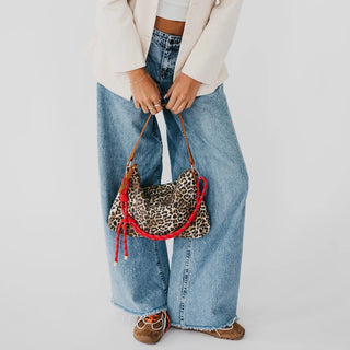 A person wearing wide-leg light blue jeans, a cream blazer, and brown shoes holds a leopard-print crescent shoulder bag with red tassels and trim against a plain white studio background.