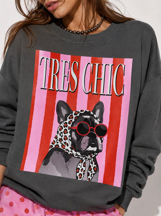A close-up of a person wearing a dark gray crewneck sweatshirt with a large graphic print. The print features pink and red vertical stripes, the text 'TRES CHIC' in white with black outlines, and an illustration of a French Bulldog wearing a leopard-print scarf and red sunglasses.