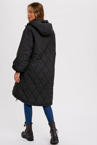 A woman wearing a long black diamond-quilted hooded coat, blue leggings, and dark brown ankle boots, shown from a rear three-quarter angle against a plain light gray studio background.