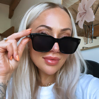 A person wearing black squared sunglasses with a white shirt on, indoors.