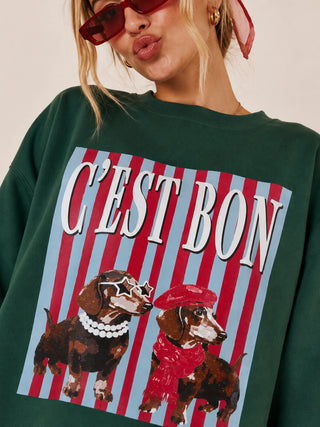 A close-up of a model wearing a dark green crewneck sweatshirt with a large graphic print. The print features the text 'C'EST BON' in white over red and light blue vertical stripes, with two dachshunds dressed in red and black accessories below.