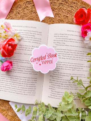 Open book with flowers and a 'Certified Book Yapper' sticker on a textured surface.