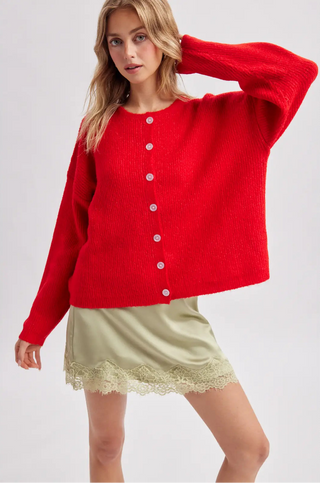 A woman modeling a bright red knit cardigan sweater with white buttons, paired with a light olive green satin skirt with a lace hem, against a light gray studio background.