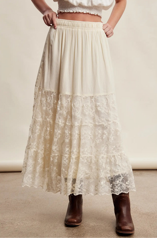 An off-white tiered maxi skirt with a solid upper section and a floral lace lower skirt, shown on a model wearing brown ankle boots against a neutral beige background.