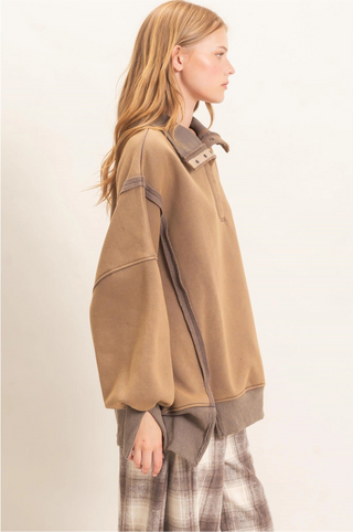 A woman shown in side profile wearing an oversized taupe-brown pullover sweatshirt with contrasting darker brown trim and a plaid skirt against a plain off-white studio background.