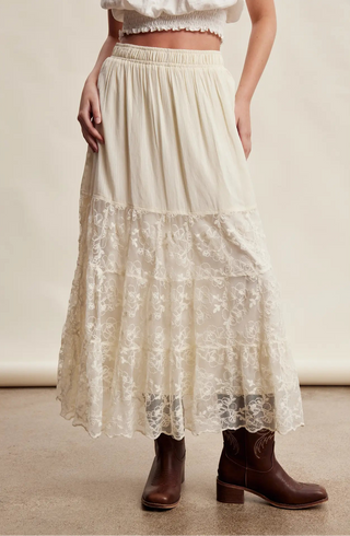 An off-white tiered maxi skirt with a floral lace overlay, worn by a model with brown leather ankle boots against a neutral beige studio background.