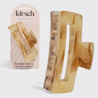 A rectangular claw hair clip made from translucent amber-brown recycled plastic, shown both on its light pink retail card and as a standalone product against a light gray background.