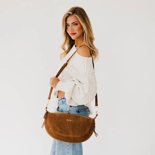 A woman wearing a white off-the-shoulder knit sweater and blue jeans carries a brown suede crescent-shaped crossbody bag with a brown leather strap against a plain white studio background.