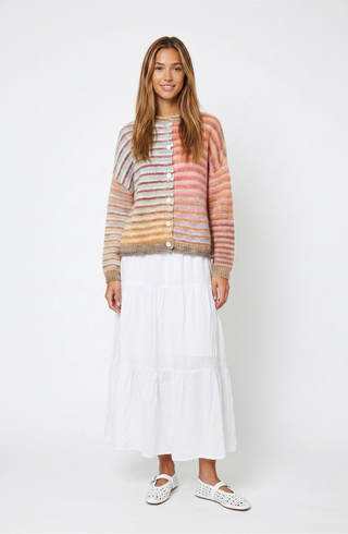 A woman stands against a light gray studio background wearing a multicolor striped knit cardigan with buttons, a long white tiered skirt, and white flat shoes.